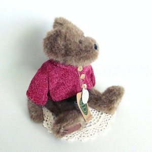 May include: A brown teddy bear wearing a magenta-coloured sweater with three gold-coloured buttons. The bear is sitting on a cream-coloured, crocheted doily. A tag is attached to the sweater.