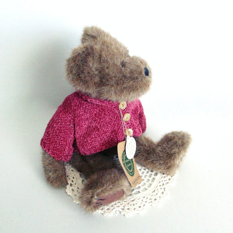 May include: A brown teddy bear wearing a magenta-coloured sweater with three gold-coloured buttons. The bear is sitting on a cream-coloured, crocheted doily. A tag is attached to the sweater.