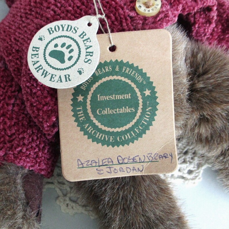 May include: A close-up of a collectible plush bear with a tag. The tag reads "Boyds Bears & Friends" and "Investment Collectables". The bear is wearing a burgundy sweater. The tag also has the name "Azalea Bosenbeary & Jordan".
