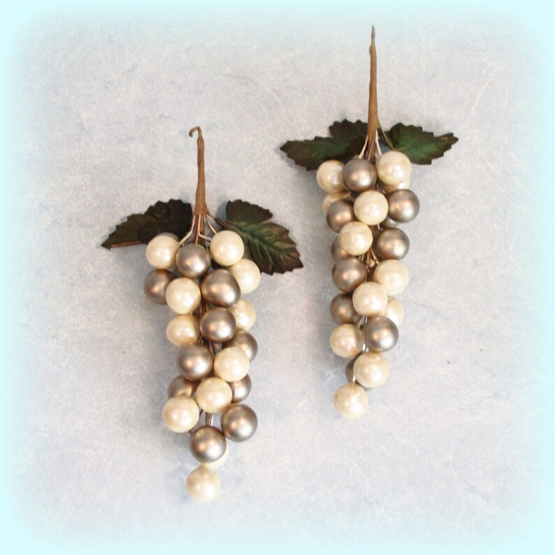 May include: Two decorative grape clusters with pearl-like beads in cream and silver. Each cluster has a brown stem and green leaves, creating a realistic appearance. Ideal for home decor or craft projects.