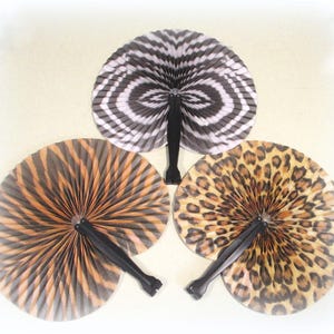 May include: Three circular hand fans with black plastic handles. One fan features a black and white zebra stripe pattern, one displays a brown and black tiger stripe design, and the third shows a tan and black leopard spot print pattern.
