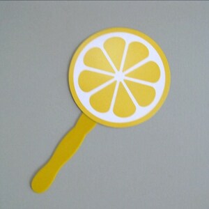 May include: A yellow hand fan with a white lemon slice design. The fan has a yellow handle.