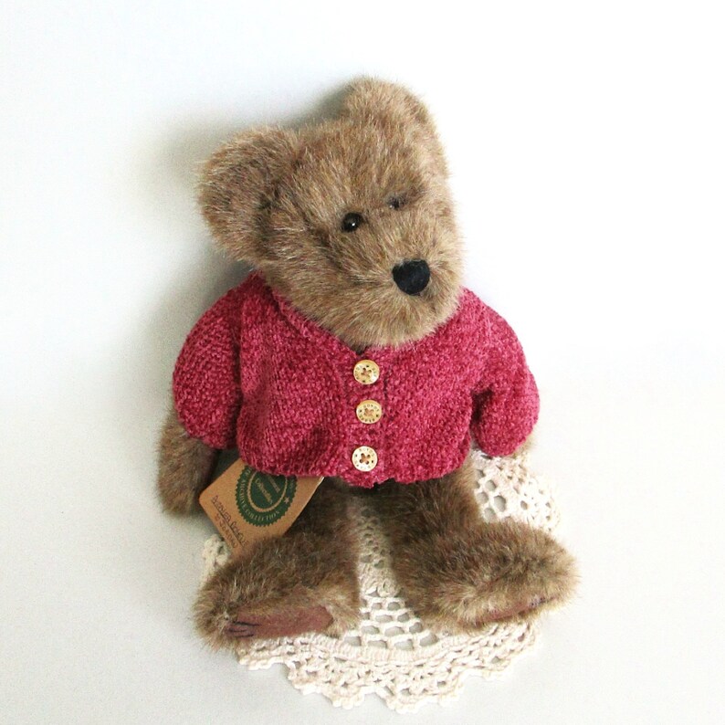 May include: A brown teddy bear wearing a crimson red cardigan with three gold buttons. The bear sits on a cream-coloured, crocheted doily. A small tag is attached to the bear's cardigan.