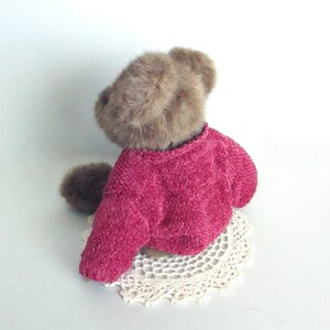 May include: A brown teddy bear wearing a bright pink sweater. The bear is sitting on a cream-coloured, crocheted doily. The fur on the bear's head and arms is a lighter brown colour.