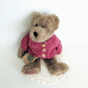 May include: A soft toy teddy bear with brown fur, wearing a magenta pink cardigan with three gold buttons. The bear sits on a white lace doily. A small tag is attached to the bear's leg.