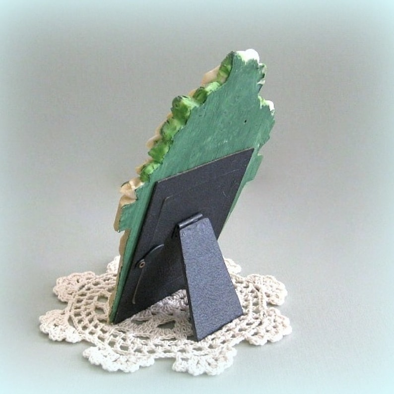 以下が含まれることがあります： 緑とクリーム色の花柄の装飾的なフォトフレーム。フレームは黒いイーゼルバックで支えられ、クリーム色のレース編みのドイリーの上に置かれています。フレームの寸法は表示されていません。