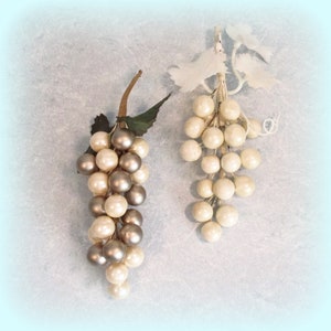 May include: Two bunches of artificial grapes. One has silver and cream-coloured orbs, green leaves and a brown stem. The other is cream-coloured with white leaves and a white stem. The background is a light blue.