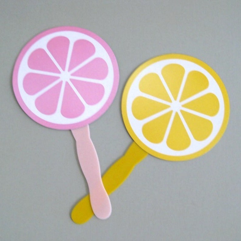 May include: Two hand fans with a lemon and a pink grapefruit design. The fans are made of plastic and have a white background with a yellow and pink citrus design.