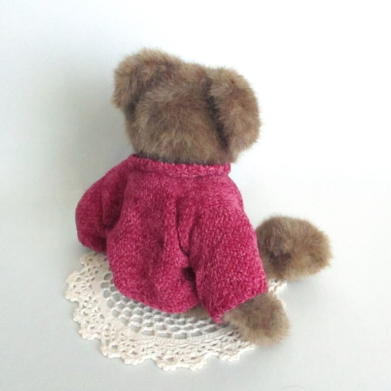 May include: A brown teddy bear wearing a magenta sweater, sitting on a cream-coloured, crocheted doily. The bear's fur has a soft, plush texture. The sweater has a textured knit appearance.