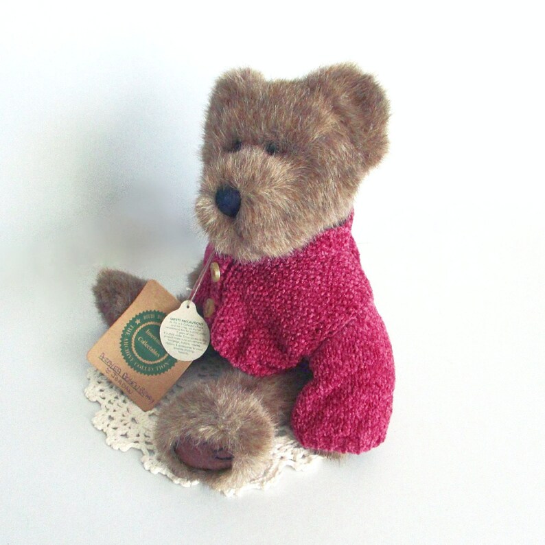 May include: A brown teddy bear wearing a pink sweater with gold buttons. The bear has a blue nose and is sitting on a white lace doily. A tag is attached to the bear's ear.