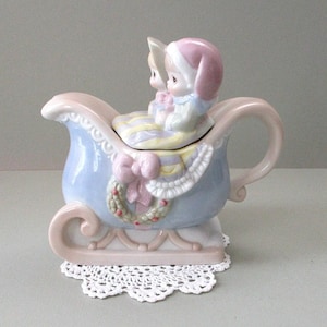 May include: A ceramic sleigh-shaped teapot featuring a light blue body with a pink handle and base. The lid is topped with two figures wearing pink and white hats, wrapped in a striped blanket. A pink bow and a wreath are sculpted on the side.