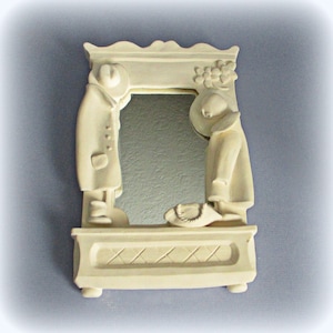 May include: A white ceramic mirror with a decorative frame featuring two figures in relief. The figures are dressed in clothing and appear to be standing in front of a mirror. The frame has a scalloped top and a base with a diamond pattern.
