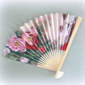 May include: A hand fan with a floral design featuring pink and red peonies on a green background. The fan is made of paper and has a wooden handle.