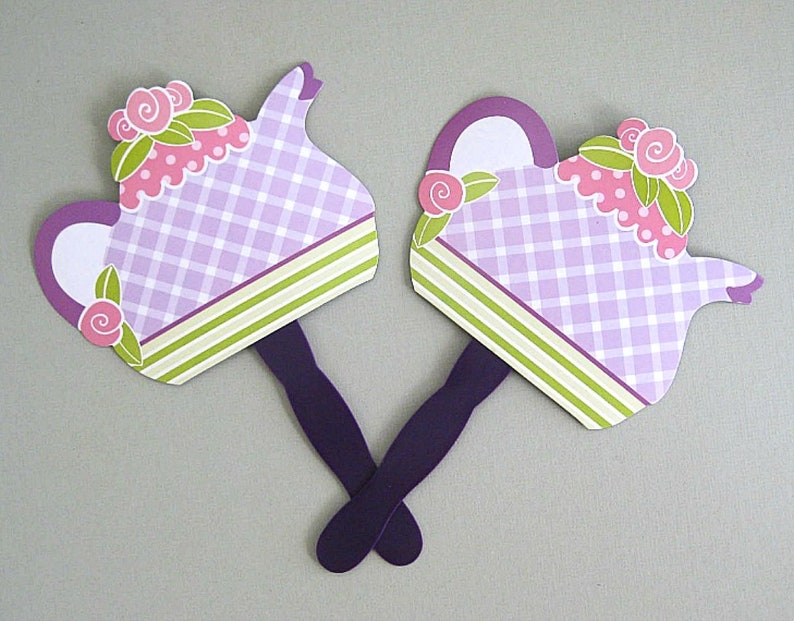 May include: Two purple and white teapot-shaped paper fans with green and pink floral accents. The fans have purple handles.