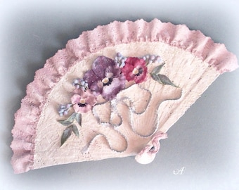 May include: A delicate, hand-painted fan with a pink lace trim and a floral design featuring purple and pink pansies. The fan is made of a textured, white material and has a delicate, feminine feel.