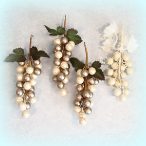 May include: Four bunches of artificial grapes. The grapes are made of pearl-like beads in white and silver, attached to brown stems with green leaves. One bunch is all white.