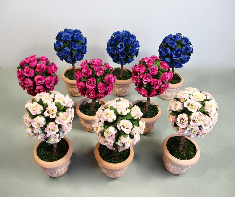 May include: Nine miniature potted topiary trees with pink, blue, and white paper roses. The trees are in terracotta pots with moss around the base.