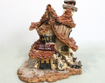 May include: A detailed miniature cottage figurine with a thatched roof, chimney, and the sign "Bearly Well Clinic." The cottage has a stone and wood exterior, with small windows and a door. The base is textured to resemble a natural landscape.