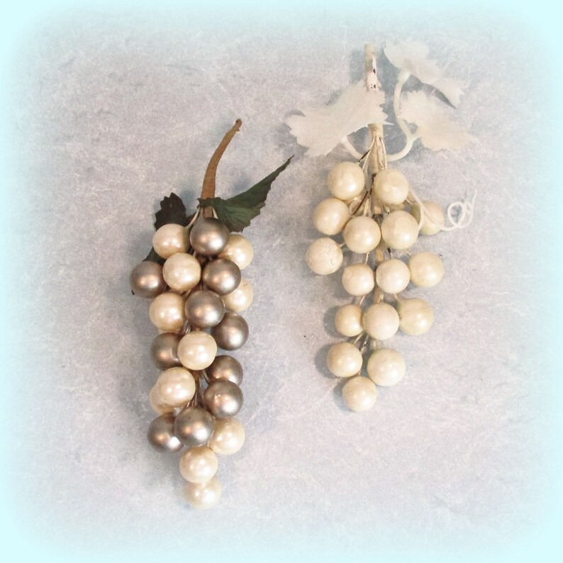 May include: Two bunches of artificial grapes. One has silver and cream-coloured orbs, green leaves, and a brown stem. The other is cream-coloured with white leaves and a white stem. The background is a light blue.