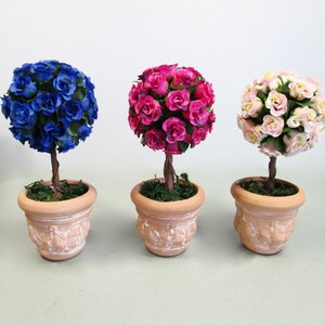 May include: Three potted topiary trees with artificial flowers. The trees are in terracotta pots with a whitewash finish. The flowers are blue, pink, and white.