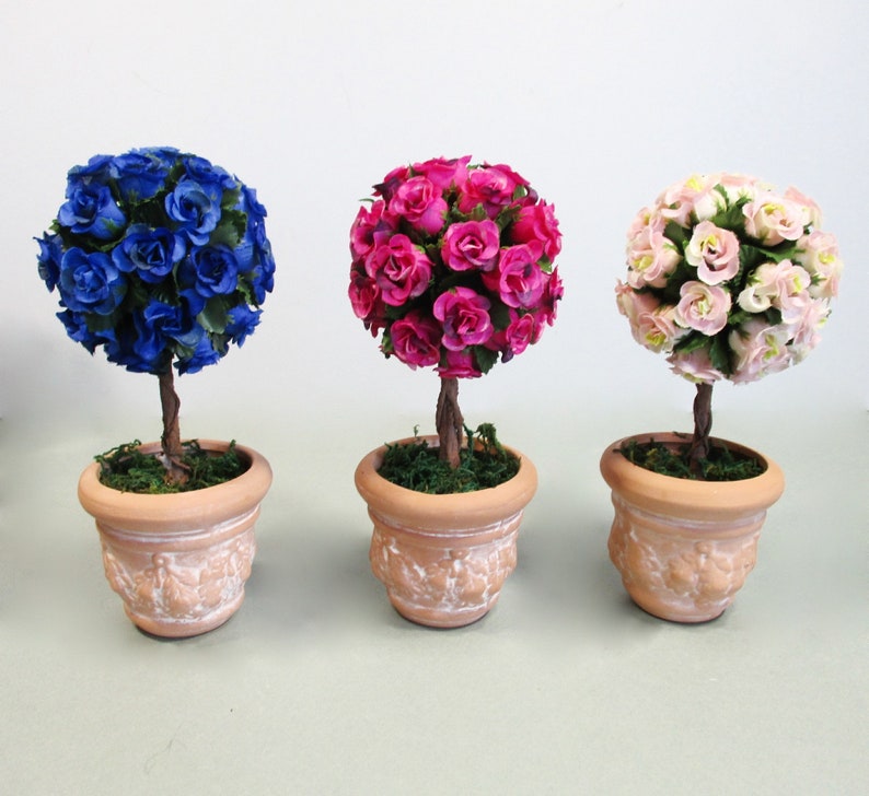 May include: Three potted topiary trees with artificial flowers. The trees are in terracotta pots with a whitewash finish. The flowers are blue, pink, and white.