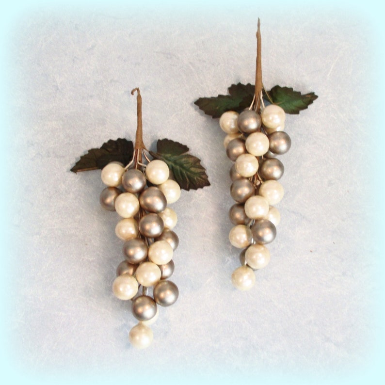 May include: Two decorative grape clusters with pearl-like beads in cream and silver. Each cluster has a brown stem and green leaves, creating a realistic appearance. Ideal for home decor or craft projects.