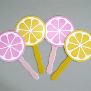 May include: Four paper hand fans with yellow and pink lemon designs. The fans have white centers and are attached to wooden sticks.
