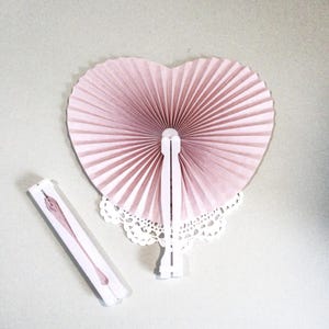 May include: A heart-shaped, pleated paper hand fan in a soft dusty pink color with a white plastic handle, accompanied by a matching rectangular box featuring a printed illustration of the fan on its exterior surface.