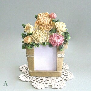 以下が含まれることがあります： クリーム、ピーチ、ピンクの花のブーケが特徴の装飾的なフォトフレーム。 フレームはバケツの形をしており、白いレースのドイリーの上に置かれています。 左下隅に文字「A」が表示されています。