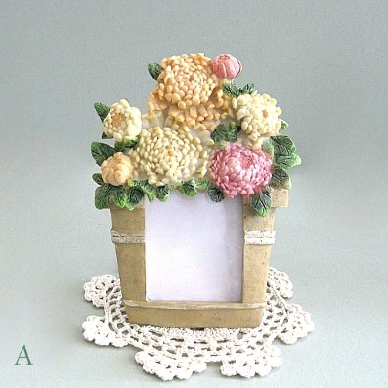 以下が含まれることがあります： クリーム、ピーチ、ピンクの花のブーケが特徴の装飾的なフォトフレーム。 フレームはバケツの形をしており、白いレースのドイリーの上に置かれています。 左下隅に文字「A」が表示されています。
