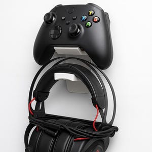 Puede incluir: Mando Xbox negro y auriculares para juegos montados en una pared blanca. El mando tiene botones y joysticks de colores. Los auriculares son negros con detalles en rojo y un logotipo en la orejera.