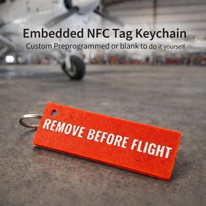 Puede incluir: Llavero rectangular de color naranja brillante con el texto blanco «REMOVE BEFORE FLIGHT». La etiqueta está unida a un llavero plateado. El fondo muestra un avión en un hangar.