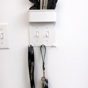 Könnte beinhalten: Ein weißer Wandorganizer mit integrierter Lichtschalterabdeckung. Er enthält schwarze Sonnenbrillen, ein Multitool und andere kleine Gegenstände. Ein Schlüsselband und ein Schlüsselring mit Schlüsseln hängen darunter. Die Wand ist weiß.