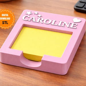 Puede incluir: Un organizador de escritorio rosa con el nombre "CAROLINE" en letras blancas, adornado con estrellas y un corazón. Contiene un bloc de notas adhesivas amarillas. La imagen también incluye un gráfico de "DESCARGA DIGITAL STL".