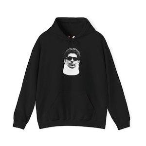 May include: Black hoodie with a front pocket and a drawstring hood. The hoodie features a black and white graphic of a person wearing sunglasses and a neck brace.
