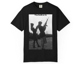 May include: Black t-shirt featuring a black and white image of two figures holding rifles, with the text "Tiocfaidh ár lá" above the image. The t-shirt is made of a soft material.