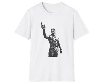 May include: A white short-sleeved t-shirt featuring a centered black and gray graphic illustration of a person with a beard and mustache, wearing a necklace and pointing one finger upward while wearing pants with a visible belt.