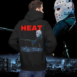 Puede incluir: Sudadera con capucha negra con la palabra "HEAT" en letras rojas en la espalda. La sudadera con capucha presenta un gráfico de una figura enmascarada sosteniendo un rifle, con un paisaje urbano en el fondo. Una segunda figura enmascarada está en el fondo.