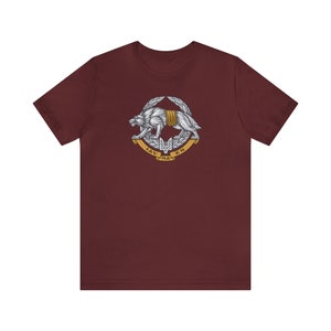 May include: A maroon t-shirt featuring a silver wolf graphic. The wolf is wearing a golden chain and is surrounded by a golden wreath. The text "IAV NA КИ" is written in gold on a banner below the wolf.