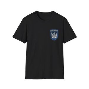 May include: Black t-shirt with a crew neck. A blue and white emblem is on the left chest, featuring text and a stylized design. The emblem appears to be a crest or logo.