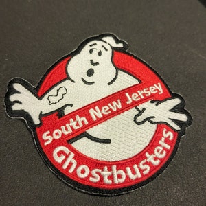 Pode incluir: Patch bordado com o logotipo dos Caça-Fantasmas. Apresenta uma figura de fantasma branco e o texto "South New Jersey Ghostbusters" em vermelho e branco. O patch tem uma borda preta e é circular.