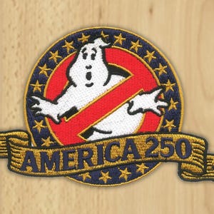 May include: A circular embroidered patch featuring a white ghost character centered over a red circle with a diagonal slash. The border is dark blue with gold stars. A gold ribbon banner across the bottom displays the text AMERICA 250 in blue.