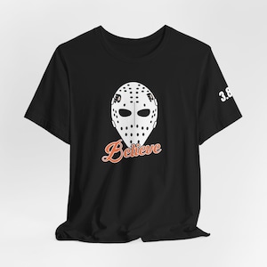 May include: Black t-shirt featuring a white hockey mask graphic with the word "Believe" in orange script. The number "3.8" is printed on the right sleeve. The design is centered on the front of the shirt.