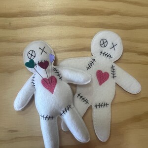 May include: Two white felt voodoo dolls with pink hearts and black stitching. One doll has green, purple, and pink hearts pinned to it.