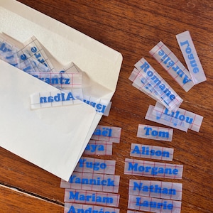 Peut inclure: Une enveloppe blanche avec un motif quadrillé sur le devant. L'enveloppe est entourée de petits autocollants rectangulaires transparents avec du texte bleu. Le texte sur les autocollants comprend des noms tels que Tom, Alison, Morgane, Nathan, Laurie et Orianne.