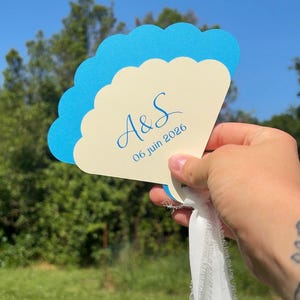 May include: A fan-shaped wedding favor with a scalloped edge, featuring a light blue and cream color scheme. The fan displays the initials "A&S" and the date "06 juin 2026". A white ribbon is attached to the base.