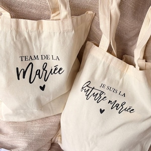 May include: Two off-white canvas tote bags with black text and heart icons. One bag reads TEAM DE LA Mariee, the other reads JE SUIS LA future mariee. Below them are pink heart-shaped sunglasses and a white sash with the text Future Mariee.