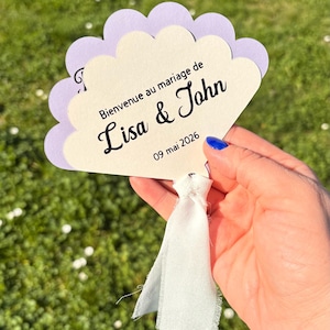 Peut inclure: Une invitation de mariage en forme d'éventail, avec des bords festonnés lavande et crème. Le texte "Bienvenue au mariage de Lisa & John" et la date "09 mai 2026" sont imprimés. Un ruban blanc est attaché.