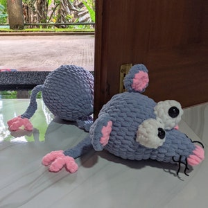 Op de afbeelding: Een handgemaakte, grijze gehaakte muisdeurstopper met roze oren en voeten, en grote witte ogen met zwarte pupillen. De muis ligt op een glanzend, lichtgekleurd oppervlak.