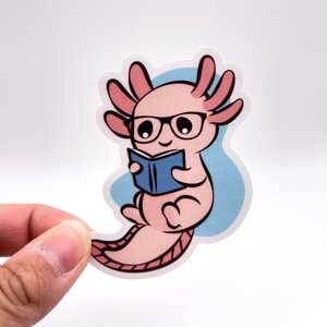 K&ouml;nnte beinhalten: Ein rosa Axolotl mit Brille, das ein blaues Buch liest. Das Axolotl sitzt auf einem hellblauen Hintergrund.
